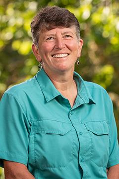 Profile photo of Colleen Schlonga, wearing a turquoise button-up shirt, in front of bright green shrubbery in the background.